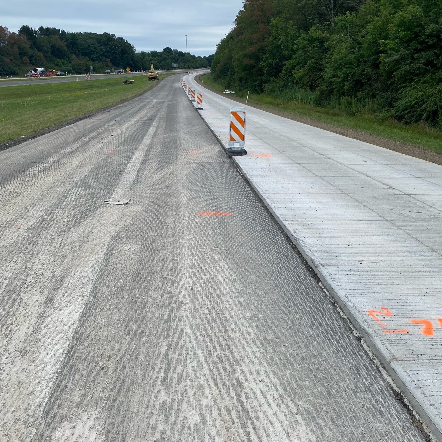 An image of a tree-lined, four-lane roadway with a grassy median between either side of the roadway with a line of orange and white work zone barricades between the completed lane and the lane be prepared for the concrete overlay